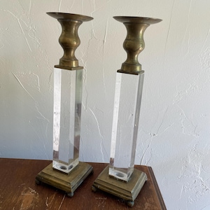 May include: Two vintage candle holders. Each has a square brass base with small feet, a clear, faceted acrylic column, and a brass candle cup. The candle holders are displayed on a wooden surface against a textured white wall.