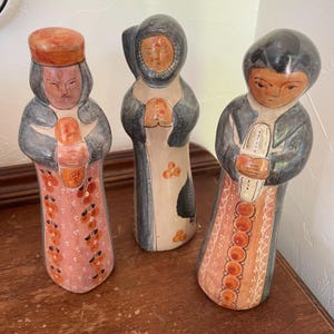 May include: Three hand-painted ceramic figurines. Each figure has a stylized face and is dressed in long robes. The colour palette includes shades of orange, pink, grey, and black. The figurines appear to be part of a religious or folk art collection.