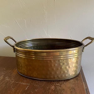 May include: A vintage brass planter with a hammered texture and two ornate handles. The oval-shaped container sits on a wooden surface. The planter is a warm gold color and appears to be well-preserved.