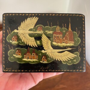 May include: A rectangular black lacquered box with a detailed painting. Two gold-colored birds fly above a landscape of churches and houses. The box has gold trim and text in the corners. The artwork is in the style of Russian lacquer miniature painting.