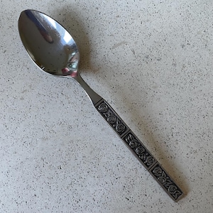 May include: A stainless steel spoon with a reflective bowl and a handle decorated with a repeating rose pattern. The spoon is set against a speckled, light-colored surface.
