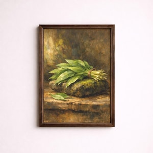 Op de afbeelding: Een ingelijst schilderij toont een bos groene bladeren, samengebonden met touw, rustend op een met mos bedekte steen. Een enkel blad ligt ernaast. Het kunstwerk heeft een donkerbruine houten lijst en een gedempt kleurenpalet.