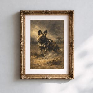 Op de afbeelding: Een ingelijste kunstdruk met een geschilderde Afrikaanse wilde hond in een naturalistische setting. Het kunstwerk is geplaatst in een sierlijke, goudkleurige lijst. De hond heeft een gevlekte vacht van zwart, bruin en wit.