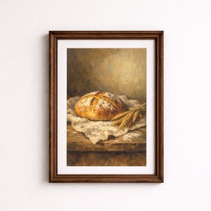 Op de afbeelding: Een ingelijst schilderij van een brood op een houten oppervlak. Het brood is goudbruin, bestrooid met bloem en ligt op een witte doek. Een bundel tarwe ligt naast het brood. De achtergrond is warm en neutraal.