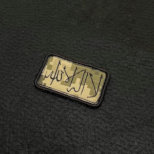 Op de afbeelding: Een rechthoekige patch met een camouflagepatroon en een zwarte rand. De patch heeft Arabisch schrift in zwart garen. De patch is waarschijnlijk een embleem of insigne.