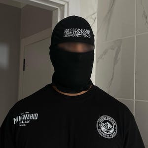 May include: Black balaclava and black t-shirt. The balaclava has white Arabic script on the forehead. The t-shirt features white text and a circular graphic with text and a mountain design.