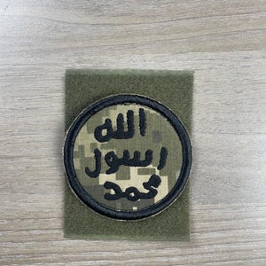 May include: A circular patch with Arabic text, featuring a black border and a camouflage pattern within the circle. The patch is set on a rectangular olive green fabric.