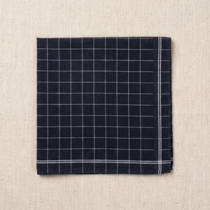 May include: A navy blue handkerchief with a white grid pattern. The square handkerchief is folded and lies flat on a beige surface. The edges are finished with a white border.