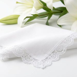 May include: A folded white handkerchief with delicate lace trim. The handkerchief is placed in front of white lilies with green stems and leaves. The lace trim adds a touch of elegance to the classic design.