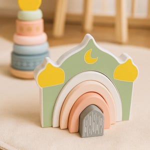 Puede incluir: Juego de apilamiento de madera con un arco en forma de mezquita y un arco iris. La mezquita es de color verde claro con detalles amarillos y una luna creciente. Un segundo juguete apilable es una torre colorida.