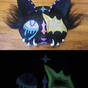 May include: A black cat mask with colourful accents, including a blue tear, pink star, and yellow and green shapes. The mask has white teeth and black fur. The mask glows in the dark, revealing the colourful accents.