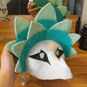 May include: A textured mask featuring a white, pointed face with black-rimmed eye openings and a vibrant teal and light green spiky crest. A small dangling charm with white and translucent beads hangs from the lower left side of the mask structure.