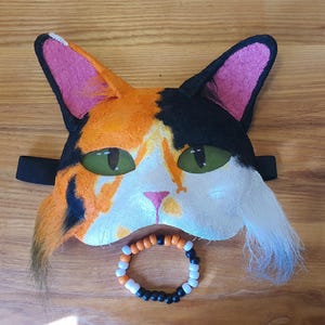 May include: A calico cat mask featuring orange, black, and white fur-like textures, pointed ears with pink inner lining, and green eyes. The mask includes a small pink nose and a black elastic strap. Below the mask sits a circular beaded bracelet.