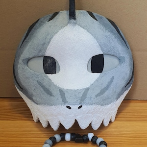 Op de afbeelding: Een grijs en wit haaienmasker met een rugvin, zwarte gaasbedekking voor de ogen en een getande onderrand, vergezeld van een ronde armband gemaakt van zwarte, witte en grijze plastic kralen in verschillende vormen.