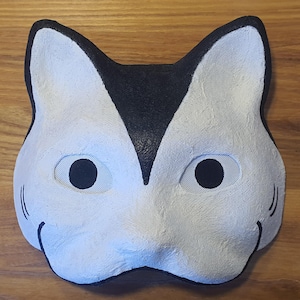 May include: A white cat mask with black detailing. The mask features mesh eye coverings and a black triangle on the forehead. The mask is made of a textured material, set against a wooden background.