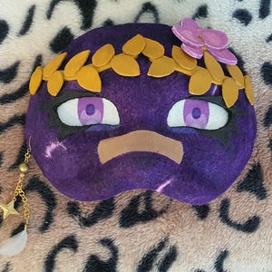 May include: A purple felt mask with large, expressive eyes, a tan mouth, and a gold leaf crown. A pink flower and a gold chain with stars and a feather dangle from the side. The mask is on a leopard print background.
