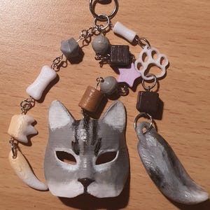 Può includere: Un portachiavi a tema gatto con una maschera di gatto grigio e bianco, un'impronta di zampa, una coda e varie perline. Il portachiavi include un moschettone per un facile attacco. La maschera di gatto ha un design dipinto dettagliato.