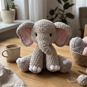 May include: A plush, crocheted elephant toy made from soft, light brown yarn. The elephant features large ears with light pink inner sections, dark safety eyes, a trunk, and white yarn accents on the tips of its four feet.