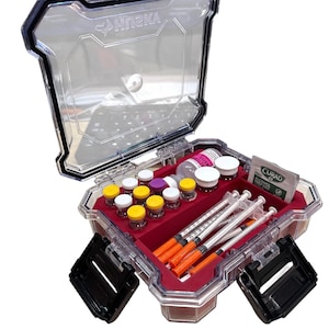 May include: A clear, hard-shell plastic storage case with black latches containing a medical supply kit. Inside, a red foam insert holds multiple small glass vials with colored caps, several syringes with orange caps, and a Curad alcohol prep pad.