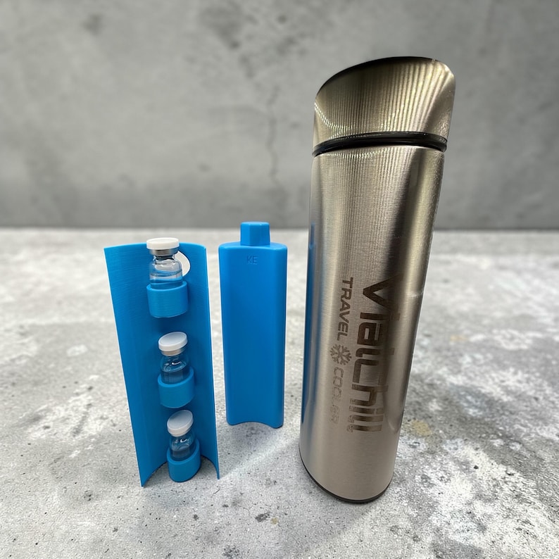 Op de afbeelding: Een roestvrijstalen reiskoeler met de tekst "VIALCHILL TRAVEL COOLER" en een zwart deksel. Een blauwe houder met vier flesjes en een blauw rechthoekig stuk liggen naast de koeler. De flesjes hebben witte doppen.