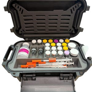 May include: A dark gray, hard-shell protective carrying case with an open lid, containing a molded insert holding multiple small glass vials with various colored caps, a larger bottle labeled Bacteriostatic Water, and several orange-capped syringes.
