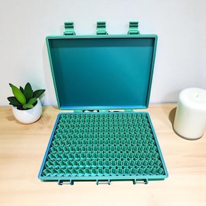 May include: An open teal-colored case with a grid of small, cylindrical compartments. The case has a matching teal interior and three latches. A small potted plant and a white candle are visible in the background.