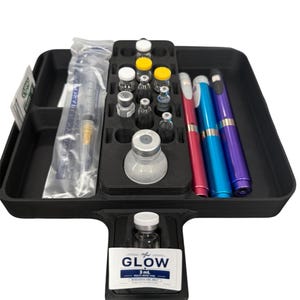 May include: A black plastic organizer tray containing a syringe in a plastic sleeve, multiple small glass vials with white and yellow caps, one large glass vial, and three metallic pens in pink, blue, and purple. One vial features a label reading GLOW.