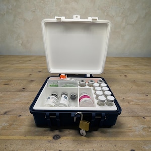 May include: An open, navy blue and white medical kit with a lock. The kit contains vials, syringes, and other medical supplies. The kit is on a wooden surface.