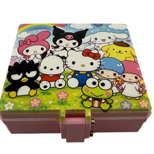 May include: A pink and white rectangular box featuring a colorful illustration of various cartoon characters, including Hello Kitty. The box has a pink base and a hinged lid with a metal clasp. The top of the box has a green grassy background.