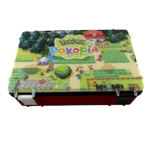 May include: A red and black case with a colorful landscape design featuring the text "Pokemon Pokepia". The case has a detailed, textured surface with a cartoonish scene of a farm, trees, and various characters.
