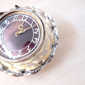 May include: A vintage, round clock with a faceted, clear crystal case. The clock face is a dark purple color with white numerals and gold-colored hands. The clock is a decorative item.