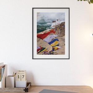 Könnte beinhalten: Gerahmte Fotografie einer Berglandschaft mit einem weißen Gebäude auf einem felsigen Hügel. Bunte Gebetsfahnen in Rot, Blau, Gelb und Weiß sind über das Gelände drapiert. Der Himmel ist bewölkt.