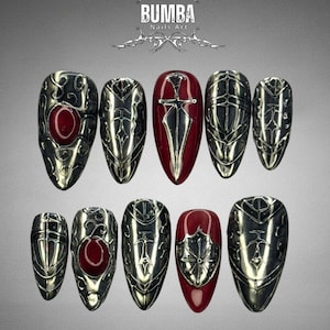 Puede incluir: Un conjunto de diez uñas artificiales con un acabado plateado metálico, con diseños intrincados y detalles en rojo. Las uñas muestran varios motivos de escudos y espadas. La marca "BUMBA Nails Art" se muestra en la parte superior.