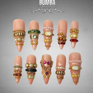 Peut inclure: Dix faux ongles, chacun orné de bijoux élaborés. Les ongles sont d'une teinte rose naturelle et présentent des ornements tels que des perles, des chaînes en or et des pierres précieuses. Le texte "BUMBA Nails Art" est visible en haut de l'image.