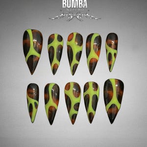 May include: A set of ten long, pointed artificial nails. Each nail features a glossy finish with a design of brown, orange, and neon green patterns. The nails are arranged on a gradient gray background. The text "BUMBA Nails Art" is at the top.
