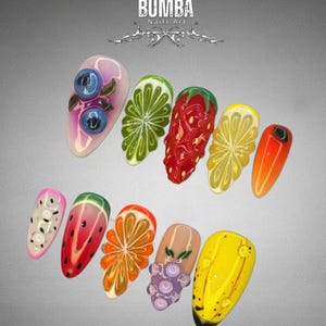 May include: A collection of ten artificial nails with fruit-themed designs. The nails feature detailed artwork of blueberries, limes, strawberries, lemons, oranges, watermelon, grapes, and a yellow banana. The text "BUMBA Nails Art" is at the top.
