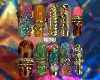 May include: A set of ten press-on nails featuring diverse designs including floral patterns, metallic gold accents, embedded colorful rhinestones, and textured finishes. The nails display various colors like orange, green, brown, and pink with the BUMBA Nails Art logo.