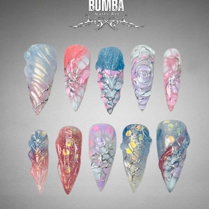 May include: A collection of ten long, pointed acrylic nails with intricate designs. Each nail features a unique combination of colors, including pink, blue, and purple, with silver accents and textured patterns. The brand name "BUMBA Nails Art" is displayed at the top.