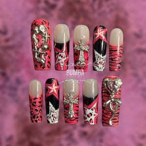 May include: A set of ten artificial nails with a variety of designs. The nails feature red, black, and white patterns, including leopard print, stars, and stripes. Some nails have silver embellishments, such as crosses and figures.