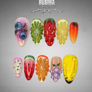 May include: A collection of ten long, almond-shaped artificial nails with vibrant fruit-themed designs. The designs include blueberries, lime, strawberry, lemon, orange, dragon fruit, watermelon, and grapes. The nails are set against a gray background, with the text "BUMBA Nails Art" at the top.