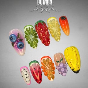 May include: A set of ten acrylic nails with fruit designs. The nails feature detailed illustrations of blueberries, limes, strawberries, lemons, oranges, watermelons, grapes, and bananas. The text "BUMBA Nails Art" is at the top.