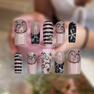 May include: A set of ten artificial nails with various designs. Some nails feature a light pink base with black and white swirls, stripes, or floral patterns. Rhinestones are added for embellishment. The word "BUMBA" is visible.