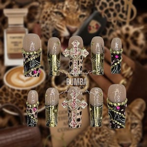 Puede incluir: Un conjunto de diez uñas artificiales con diseño de estampado de leopardo, detalles dorados y adornos brillantes. Algunas uñas presentan un diseño de cruz con gemas rosas. La marca "BUMBA Nails Art" es visible.