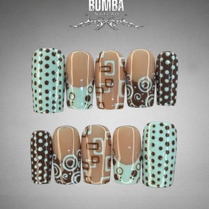 May include: A collection of ten false nails with diverse designs. The nails display light brown, teal, and dark brown hues. Some nails are adorned with polka dots, while others feature geometric and swirl patterns. The text "BUMBA Nails Art" is visible.