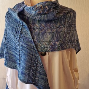 May include: A hand-knitted triangular shawl in variegated shades of blue, featuring a textured stitch pattern with vertical columns of lace detailing. The yarn displays a subtle color gradient ranging from light sky blue to deep navy tones.