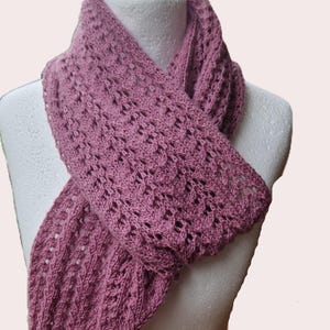 May include: A mauve-colored, open-weave scarf is draped over a mannequin. The scarf has a delicate, lacy pattern and a fringed edge. The knitted scarf is made of a soft, lightweight material, perfect for adding a touch of elegance to any outfit.