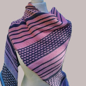 Puede incluir: Un chal de punto en tonos rosa, azul y morado, expuesto sobre un maniquí. El chal presenta un diseño a rayas y punteado. El degradado de color pasa del azul al rosa. El chal está drapeado sobre los hombros.