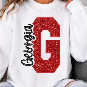 Può includere: Una felpa bianca a girocollo con una grande lettera G rossa glitterata con il centro bianco. A sinistra della lettera, la parola Georgia è scritta in un carattere corsivo nero. Il design è centrato sulla parte anteriore del capo.