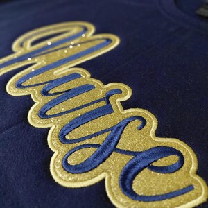 May include: Navy blue t-shirt with a gold glitter and blue embroidered "Muse" design. The word is in a cursive font with a gold glitter outline and blue embroidered letters. Small, sparkly embellishments are scattered across the design.