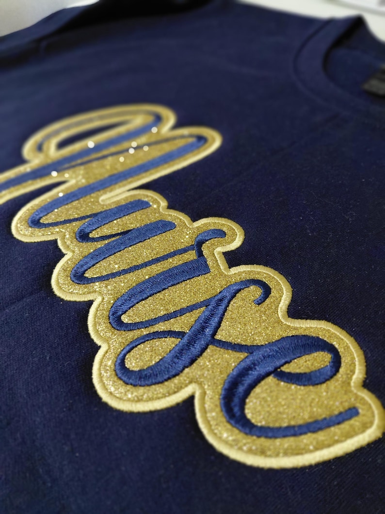 May include: Navy blue t-shirt with a gold glitter and blue embroidered "Muse" design. The word is in a cursive font with a gold glitter outline and blue embroidered letters. Small, sparkly embellishments are scattered across the design.